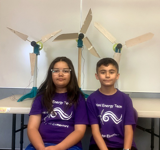 Two 4th grade boys sitting side-by-side on chairs wearing t-shirts that say, "Wind Energy Team on the top and Burlington Elementary on the bottom. Their winning turbine project is sitting on the table behind them. 