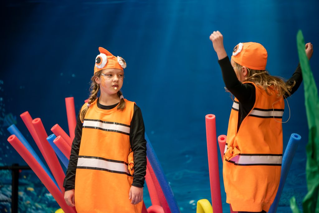 Two female elementary students on a stage dressed as clown fish. They are facing each other. The backdrop makes it look like they're under water. 