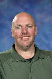 A smiling man with a shaved head, wearing a green shirt, stands in front of a blurred blue background.