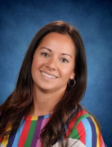 Smiling woman with long brown hair, wearing a colorful striped shirt, against a blue background.